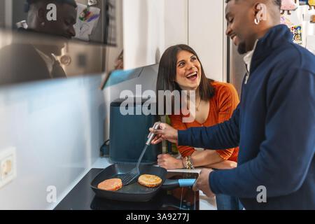 Cheerful multiethnic couple laughing and having fun while preparing burgers together on the stovetop in their modern kitchen Stock Photo