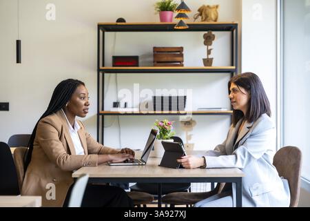 Two businesswomen are having a business meeting in a hotel lobby, working on their laptops and digital tablets Stock Photo