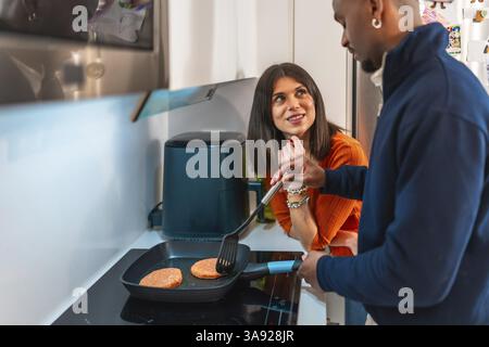 Happy multiethnic couple preparing burgers in a pan in their modern kitchen, enjoying their time together Stock Photo