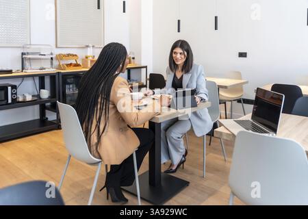 Two businesswomen enjoying breakfast and discussing work using a digital tablet in a modern hotel restaurant Stock Photo