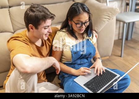 Young multiethnic couple sitting on floor and using laptop, browsing internet and smiling in cozy living room Stock Photo