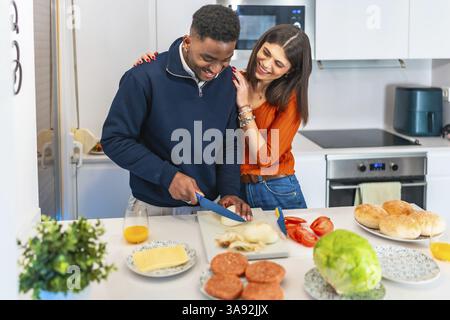 Young multiethnic couple preparing burgers, cutting onion and having fun in a modern kitchen Stock Photo