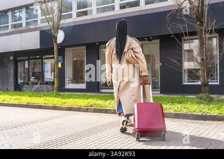 Businesswoman pulling red trolley suitcase while walking on a sidewalk towards hotel entrance during a business trip Stock Photo