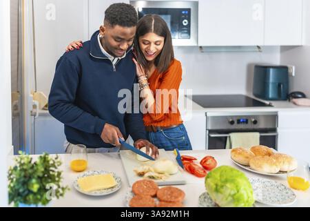 Young multiethnic couple preparing burgers together in their modern kitchen, enjoying their quality time Stock Photo