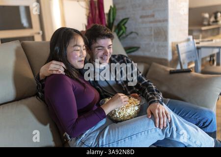 Asian woman and caucasian man enjoying leisure time at home, sitting comfortably on a sofa, sharing a bowl of popcorn in a cozy living room Stock Photo