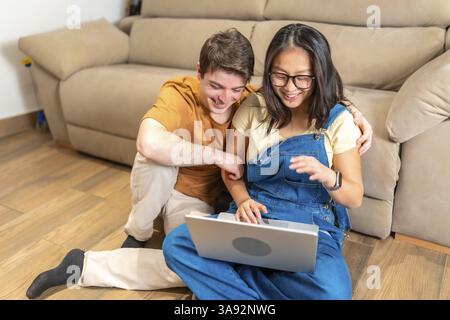 Happy multiethnic couple sitting on the floor and using laptop, surfing the web and having fun Stock Photo