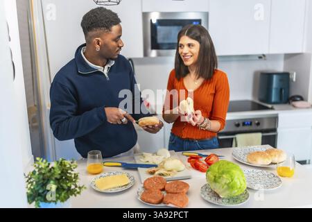 Young multiethnic couple preparing burgers in a modern kitchen, enjoying their time together Stock Photo