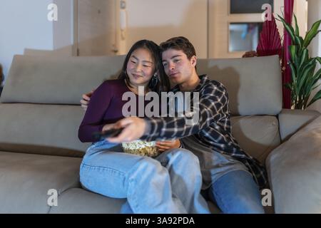 Asian woman and caucasian man relaxing on a couch, sharing popcorn and watching tv in a cozy living room setting Stock Photo