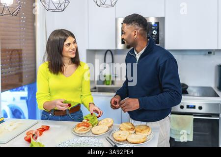 Young multiethnic couple having fun while preparing burgers for dinner in a modern kitchen Stock Photo