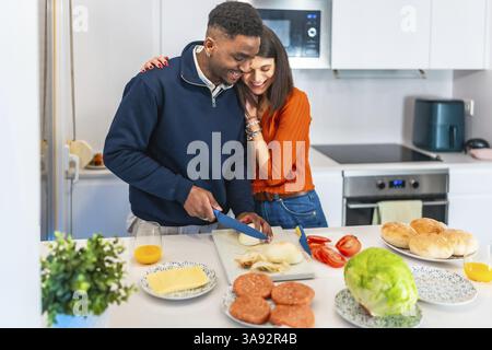 Young multiethnic couple hugging and slicing onions, preparing burgers for dinner in a modern white kitchen Stock Photo