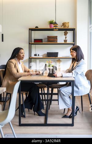 Two businesswomen are working together on their laptops and tablets in a hotel lobby, discussing business strategies Stock Photo