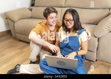 Young multiethnic couple sitting on the floor and using a laptop, browsing internet or working from home Stock Photo