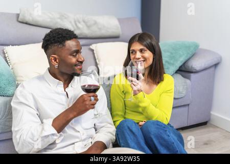Diverse couple relaxing at home, sharing wine and conversation on a cozy sofa, creating a warm and inviting atmosphere Stock Photo