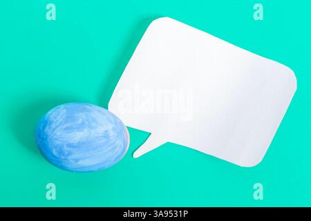 A painted blue egg placed next to a white speech bubble on a vibrant turquoise surface represents themes of communication and Easter celebration. Stock Photo