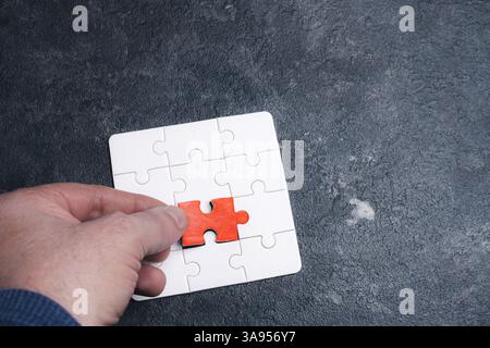 A person is holding a red puzzle piece in front of a white puzzle piece. The red piece is missing, and the person is trying to fit it into the puzzle Stock Photo