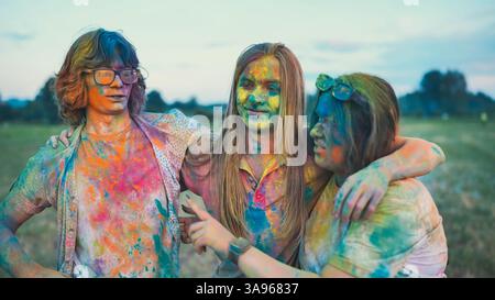 Three school friends are hugging and grinning, adorned with vibrant holi powder paint, at an outdoor summer celebration Stock Photo