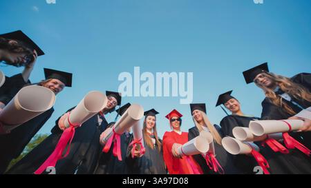 Students proudly displaying their diplomas in graduation gowns under a clear blue sky Stock Photo