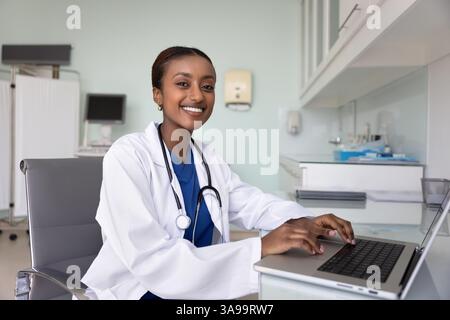 Happy beautiful African doctor woman typing on laptop at workplace Stock Photo