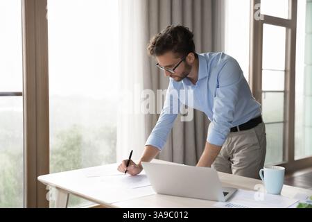 Young man standing by table review paper documents using laptop Stock Photo