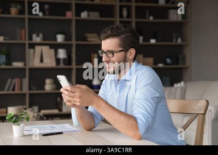 Smiling gen z male freelancer using phone at home office Stock Photo
