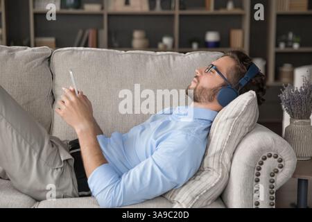 Young man wearing wireless headphones relaxing on couch holding cellphone Stock Photo