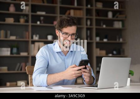 Smiling young man sit by notebook interact with smartphone app Stock Photo
