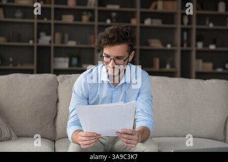 Happy smiling young man sitting on couch reading paper documents Stock Photo