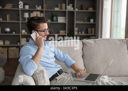 Confident young man speak on phone using notebook on sofa Stock Photo