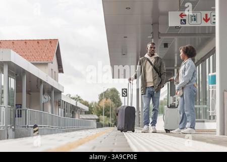 Young African American man and woman waiting for a train on the railway station platform, talking about their adventure trip. Stock Photo