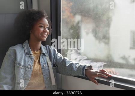 African American young woman traveling by train, enjoying her trip while looking at beautiful landscapes through the window. Stock Photo