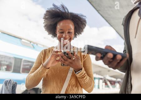 Smiling African American tourist couple with luggage using smartphone while waiting for a train on a railway station platform. Stock Photo