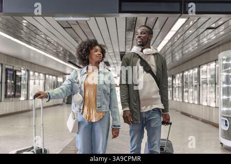Two travelers, an African American couple pulling their wheel suitcases along a train station platform. Transport, and travel concepts. Stock Photo