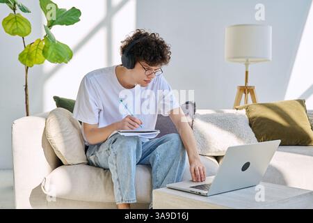 Young student guy in headphones sitting at home on with laptop taking notes Stock Photo