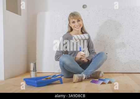 Happy smiling female painter sitting on wooden floor and holding paintroller Stock Photo