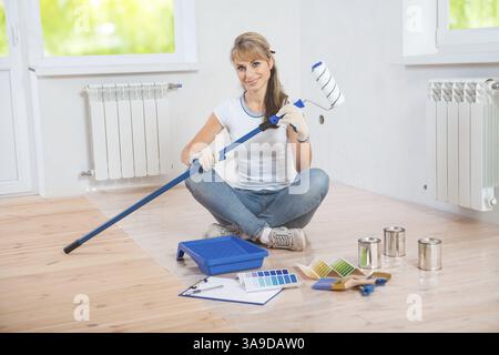 Beautiful female painter sitting on wooden floor with paint tools and holding paintroller and looking at camera Stock Photo