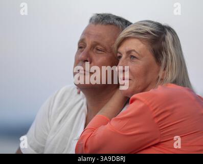A middle aged couple, a man in a white shirt and a woman in an orange one, are sitting close to each other and looking into the distance with gentle s Stock Photo