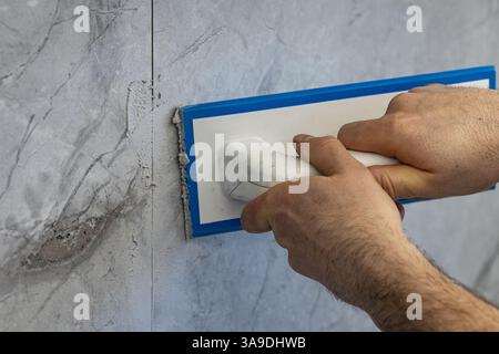 Professional worker applying grout on ceramic tile wall with a rubber float tool during renovation process. Home improvement, construction and tiling Stock Photo