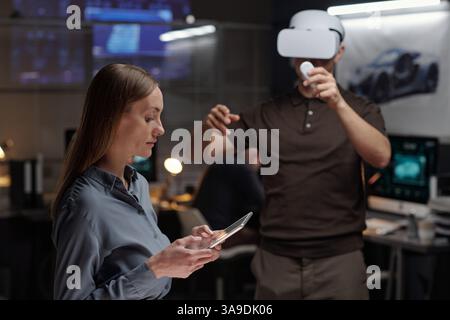 Serious female designer with tablet looking through graphic data on screen while her colleague making virtual presentation Stock Photo