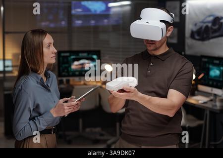 Young serious woman with tablet looking at male colleague with VR headset making presentation of new car model Stock Photo
