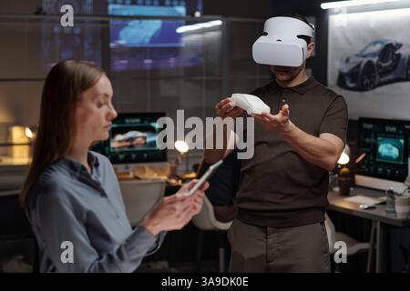 Young male engineer in VR headset holding car model on hands while standing in front of female colleague using tablet Stock Photo