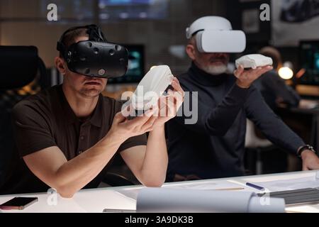 Young and mature male designers wearing VR headsets sitting by workplace in office and introducing new model of car Stock Photo