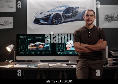 Happy young male car designer crossing his arms by chest and looking at you while standing by desktop computers in office Stock Photo