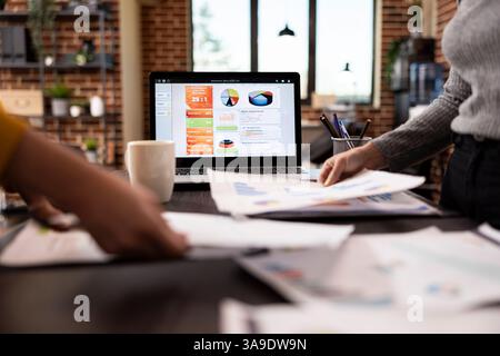 Selective focus on laptop displaying business analytics as hands of female individuals hold documents at desk. Closeup of people grasping papers over a table as digital device shows data graphs. Stock Photo
