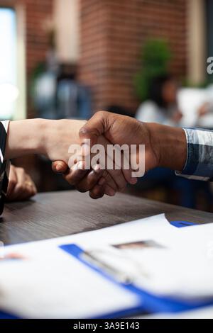 Closeup of multiethnic individuals handshaking over the office desk during job interview meeting. Selective focus on hands of diverse people doing a greeting gesture in brick wall workspace. Stock Photo
