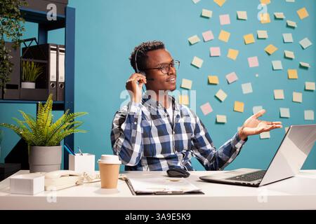 Telemarketer sitting at desk and assisting a client over the phone about application documentation. Company consultant using his laptop and handling a call, providing assistance to the employee. Stock Photo