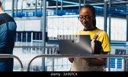 Smart factory engineer oversees operation of robotic arms and conveyor systems on facility floor. Industry 4.0 plant technician monitors system performance and troubleshoots issues, camera A Stock Photo
