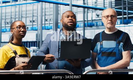 Smart factory manager teaching staff members to manage robotic arms using notebook. Supervisor overseeing industry 4.0 industrial plant, controlling automated machinery laptop, camera A Stock Photo