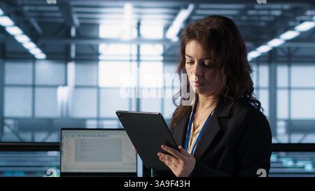 Technician in data center with tablet integrating AI driven automation tools, running predictive maintenance routines. Woman ensuring continuous uptime of server room, managing data flows, camera A Stock Photo