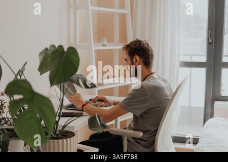 A man works mindfully at a laptop in a calm, plant filled home office. Perfect image for soft productivity, mental wellness, and remote work themes  Stock Photo