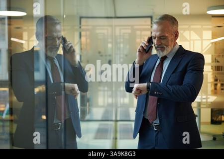 Mature Caucasian man checking time on smartwatch while talking on phone, standing in modern office with panoramic windows Stock Photo